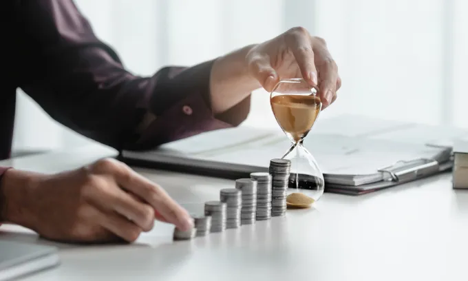 A pair of hands rest on the shortest of multiple piles of coins and an hour glass.