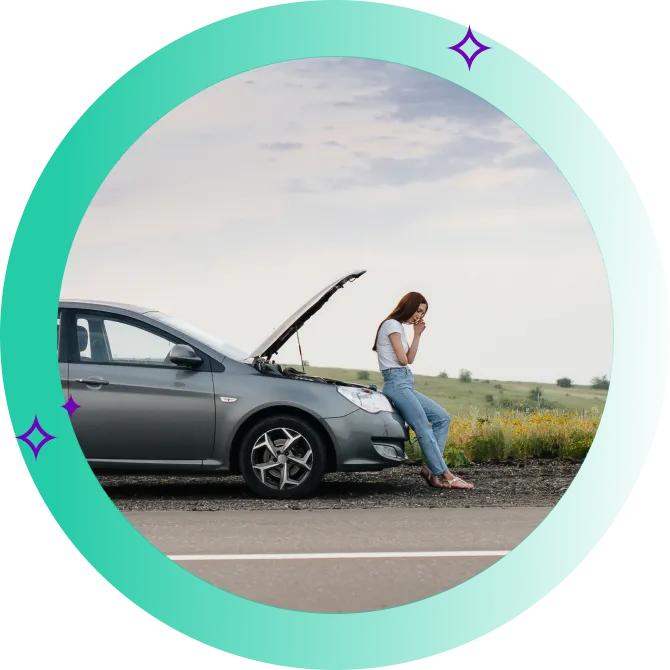 A driver calling for roadside assistance, seated against the open hood of their car