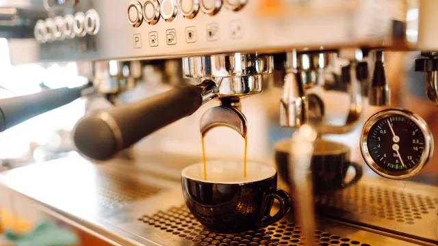 Experienced barista prepares freshly brewed coffee using professional coffee machine. In cozy coffee shop, espresso is poured from espresso machine into black cup. Small business, beverage concept.