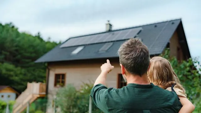 Rear view of dad holding his little girl in arms and showing at their house with installed solar panels.