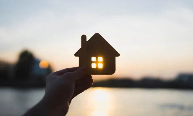 A person holds a wooden cut out of a house in front of the setting sun.