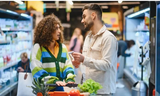 Young couple looking for items, talking and pushing shopping cart filled with fresh produce in a supermarket aisle