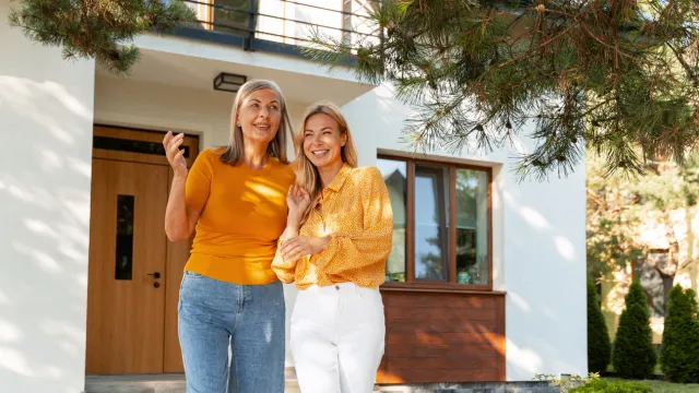 A mother and her adult daughter stand in front of a house smiling.