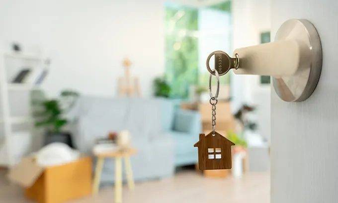 Key in lock with home-shaped keyring, door open on blurry living-room scene