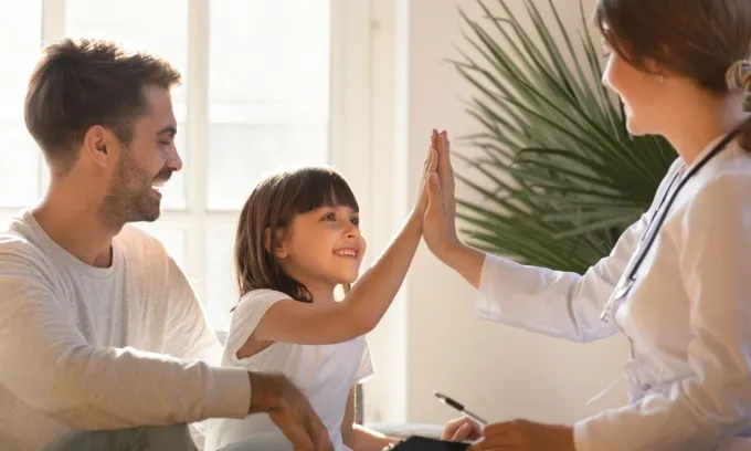 A father sits with his daughter in a doctor's office, as she high-fives the doctor.