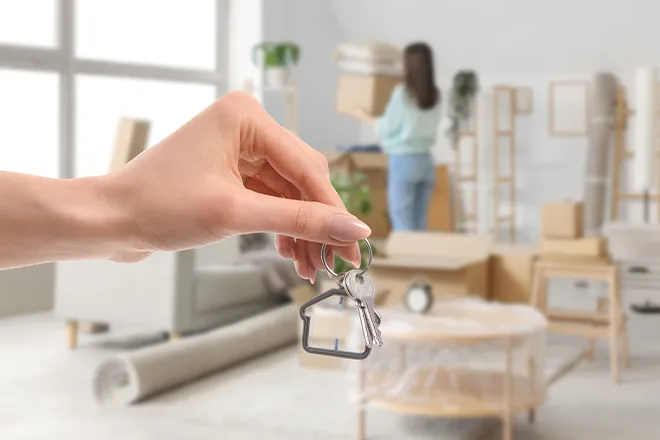 Hand holding key against backdrop of woman unpacking in new home.