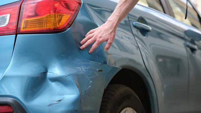 A driver runs their hand over the damaged rear of their blue car.