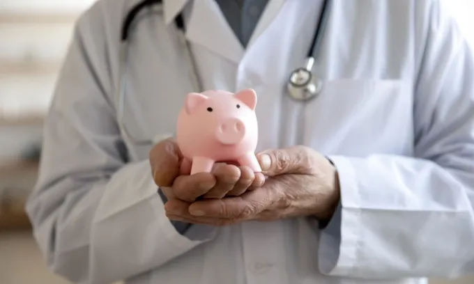 An elderly male doctor holds a pink piggy bank in his hands