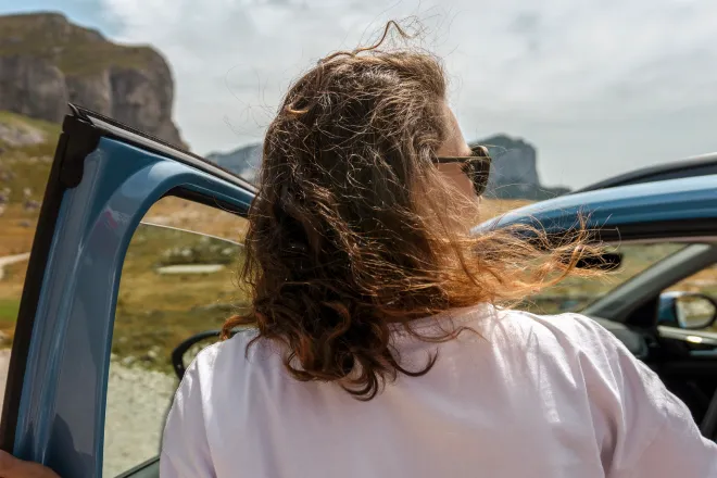 A woman about to enter her car by a scenic mountain road, her hair billowing in the wind.