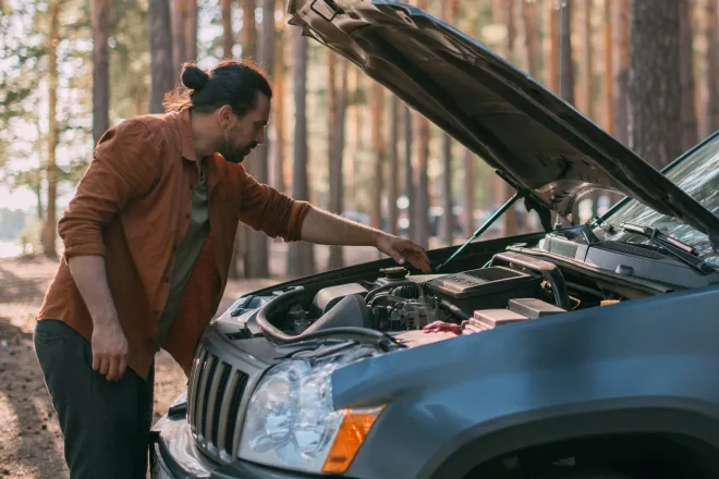 A man checking under his car bonnet by the side of a woodland road.
