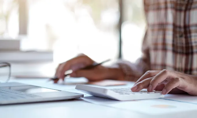 Close up of man holding pen working on calculator in front of laptop.