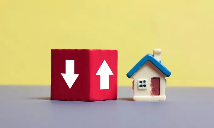 A red block with two white arrows on different sides, one pointing up and another pointing down, sits next to a small figure of a house. 