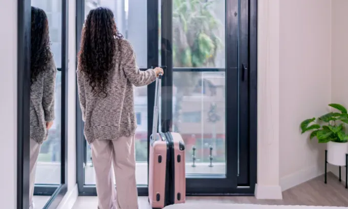 Digital nomad with rolling suitcase opening balcony door in airbnb apartment, enjoying city view
