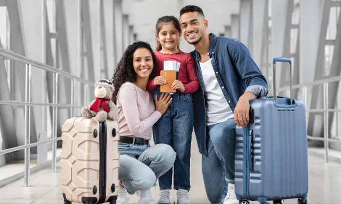 Two parents hold their suitcases while crouching next to their child who holds the passports and boarding passes.