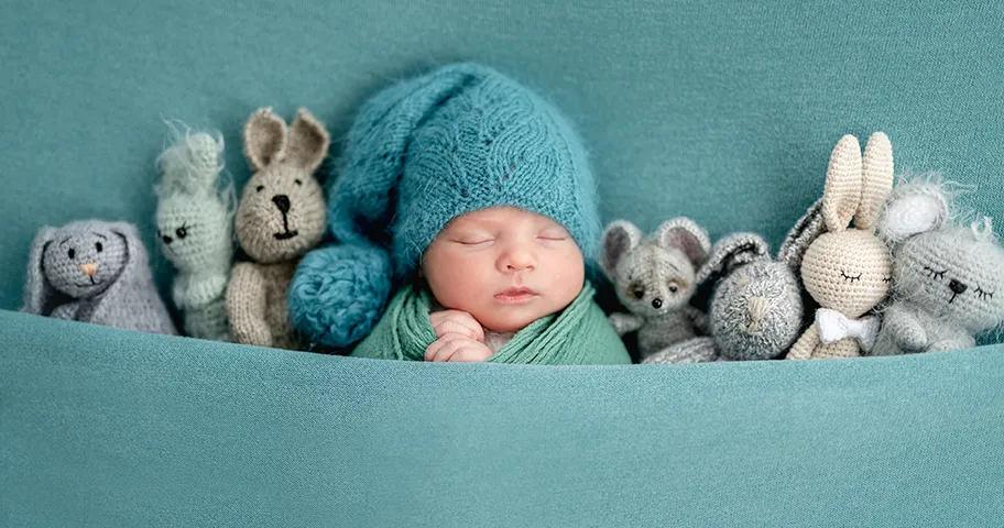 Newborn baby in bobble hat tucked up in bed with multiple knitted toys.