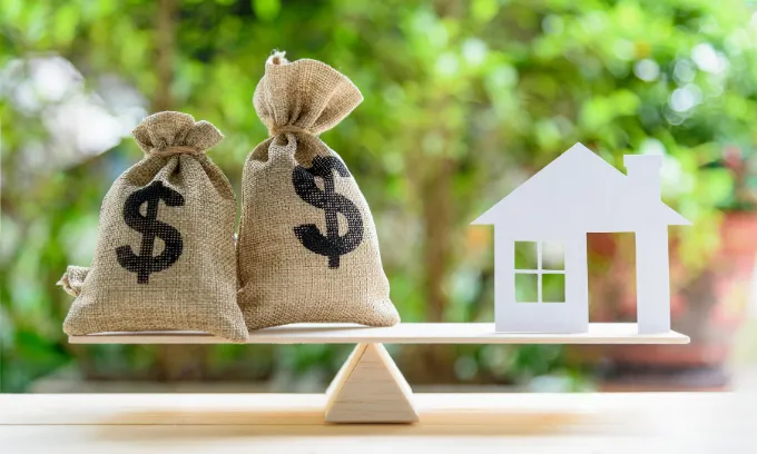 A wooden scale with two money bags on the left end and a paper house on the right.