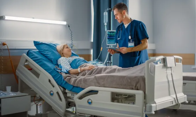 A male doctor converses with a woman who lies in a hospital bed.
