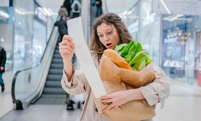 A shocked woman looks at her grocery shopping receipt while juggling her groceries.