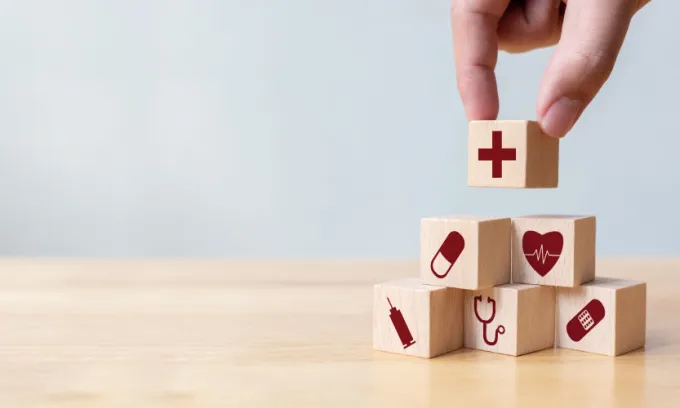 A hand places a wooden block with a red cross on it on top of a bunch of other wooden blocks with health-related images on them.