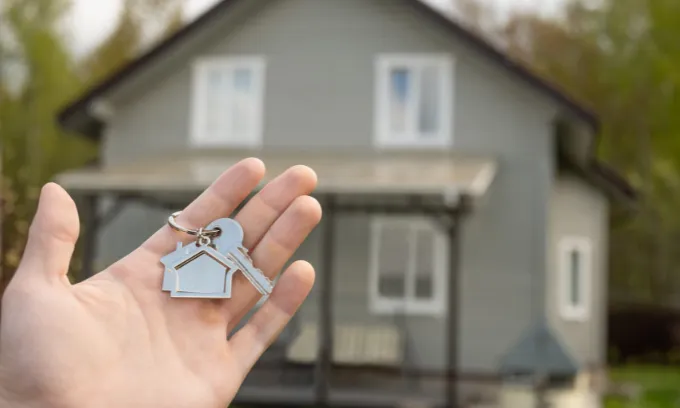 Man hand holds house-shaped key in front of newly purchased home.