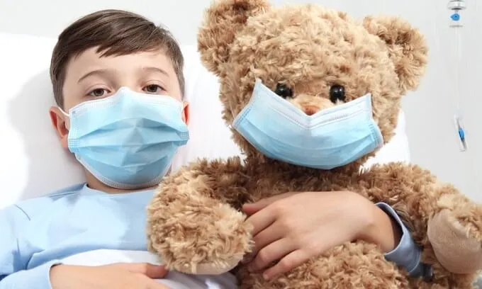A young child holds their teddy bear, both wearing blue surgical masks